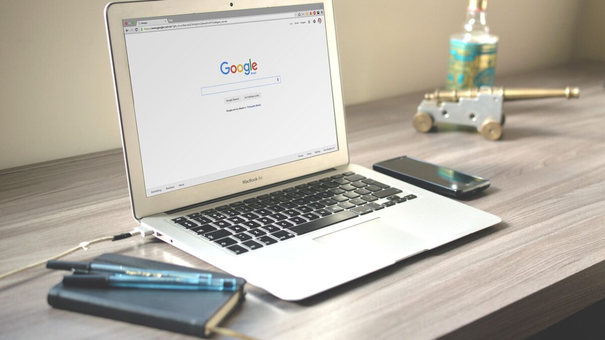 Un MacBook Air ouvert sur la page d'accueil de Google, posé sur un bureau en bois avec un smartphone, un carnet, des stylos et une petite bouteille.