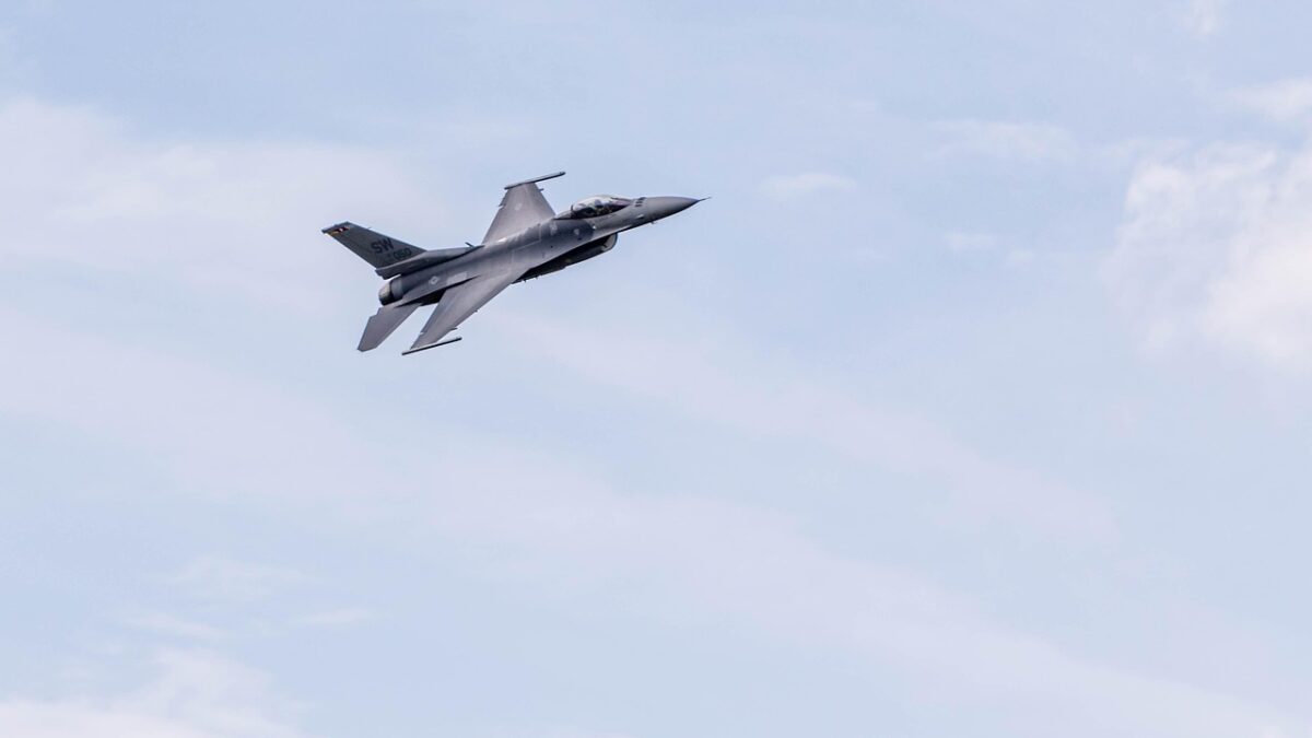 Un avion de chasse gris F-16 en vol, en virage, dans un ciel bleu nuageux, avec des marquages de queue peu lisibles.