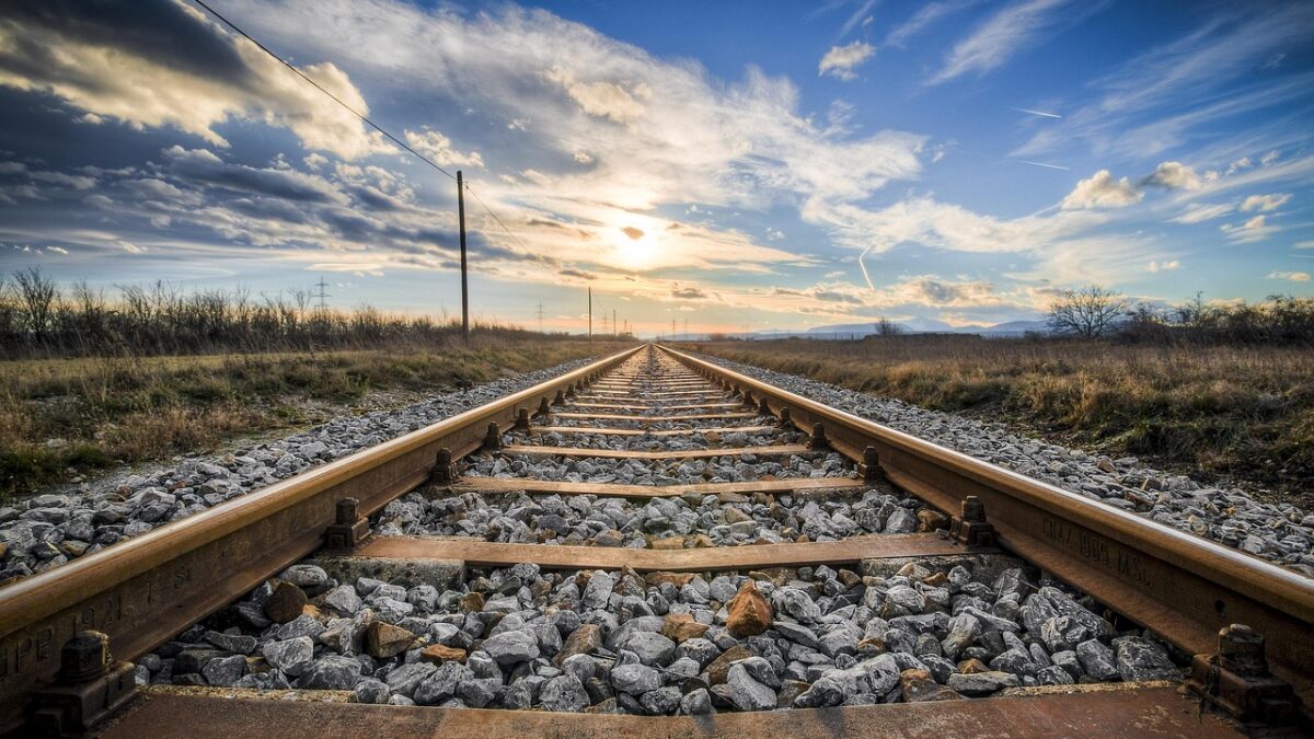 Voie ferrée en perspective, rails rouillés sur ballast de gravier, avec un poteau électrique et une caténaire à gauche, une prairie sèche et un coucher de soleil sous un ciel nuageux.