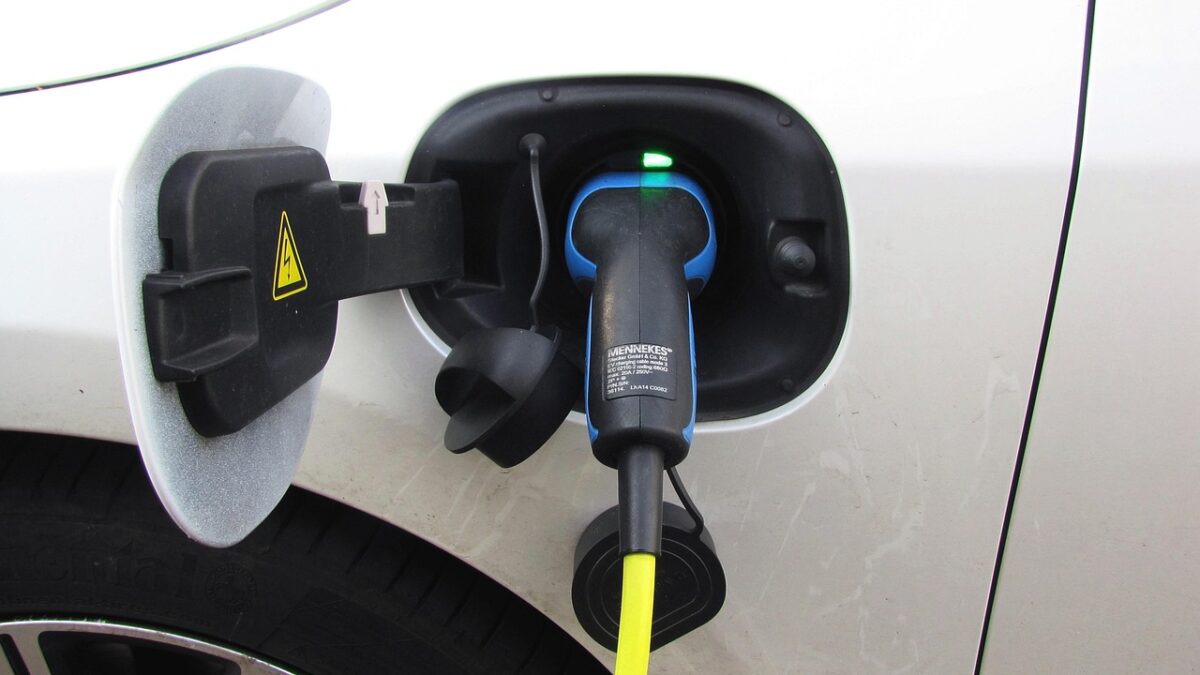 Gros plan sur la prise de recharge d'une voiture électrique blanche, avec un connecteur Mennekes bleu et noir branché, un câble jaune et un voyant vert allumé. Un pneu est visible à gauche.