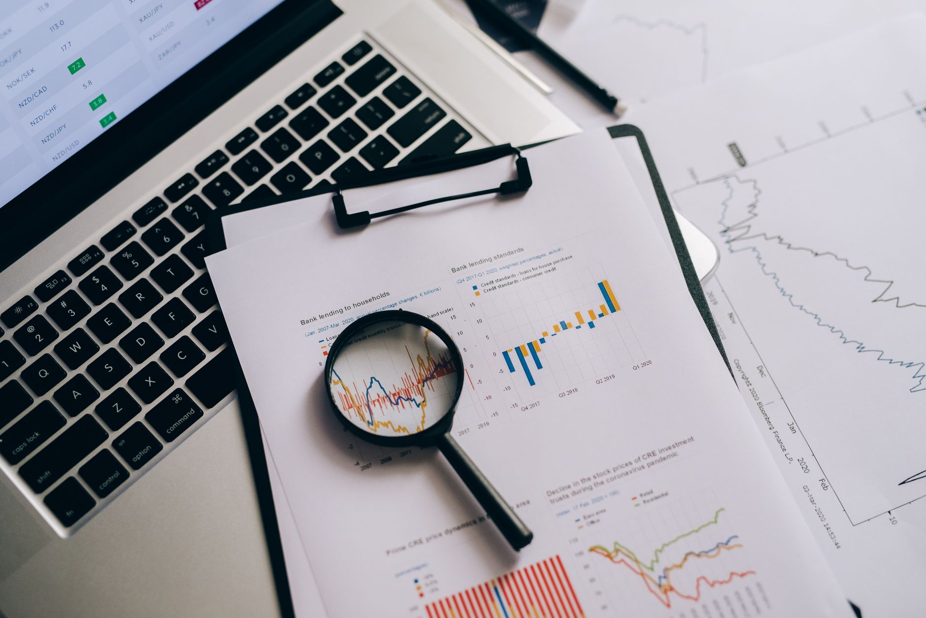 Desk with a laptop, a clipboard of printed financial charts and a magnifying glass resting on a line graph.