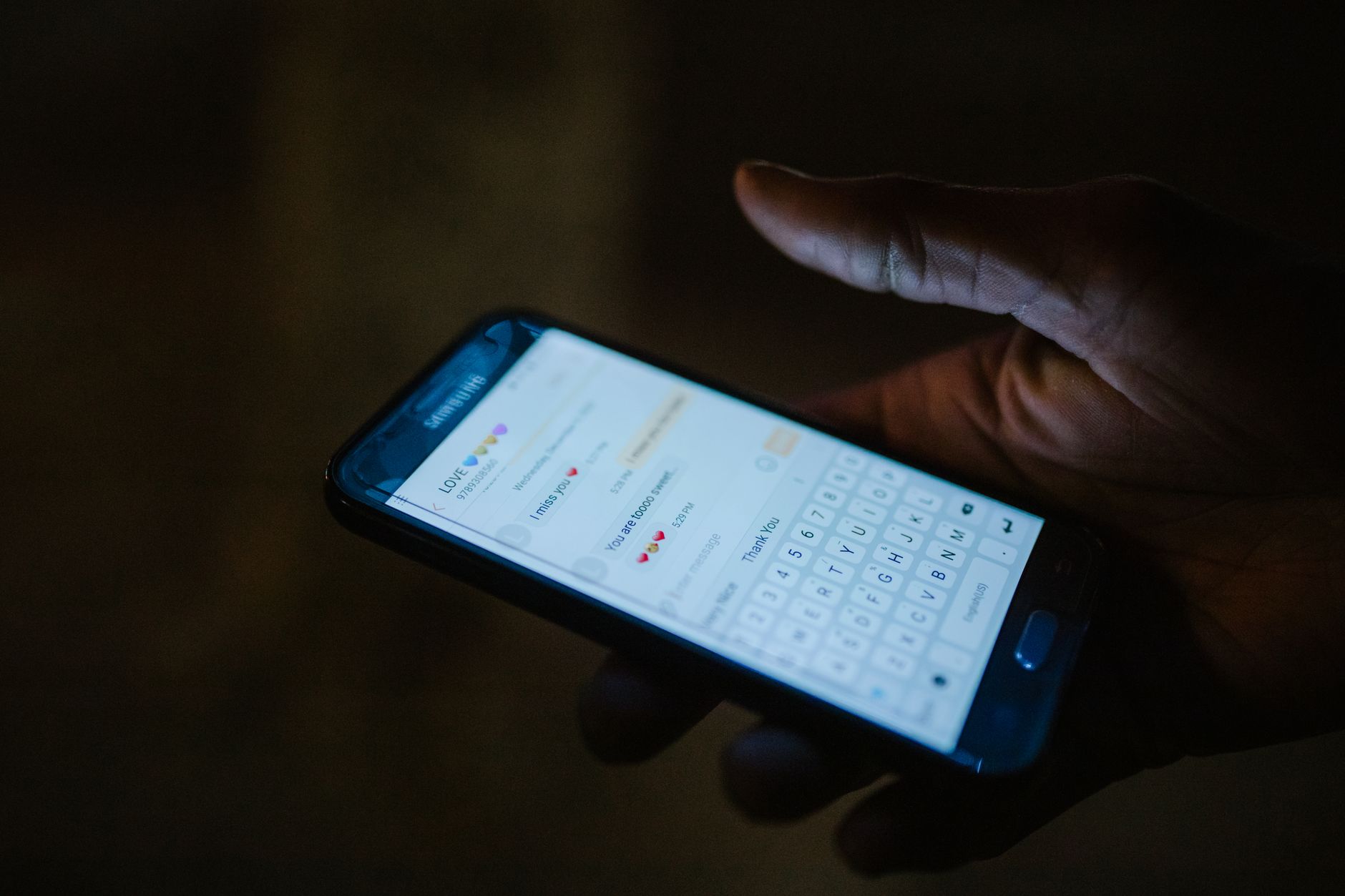 A hand holds a smartphone in the dark, its lit screen showing a text conversation and an on-screen keyboard.