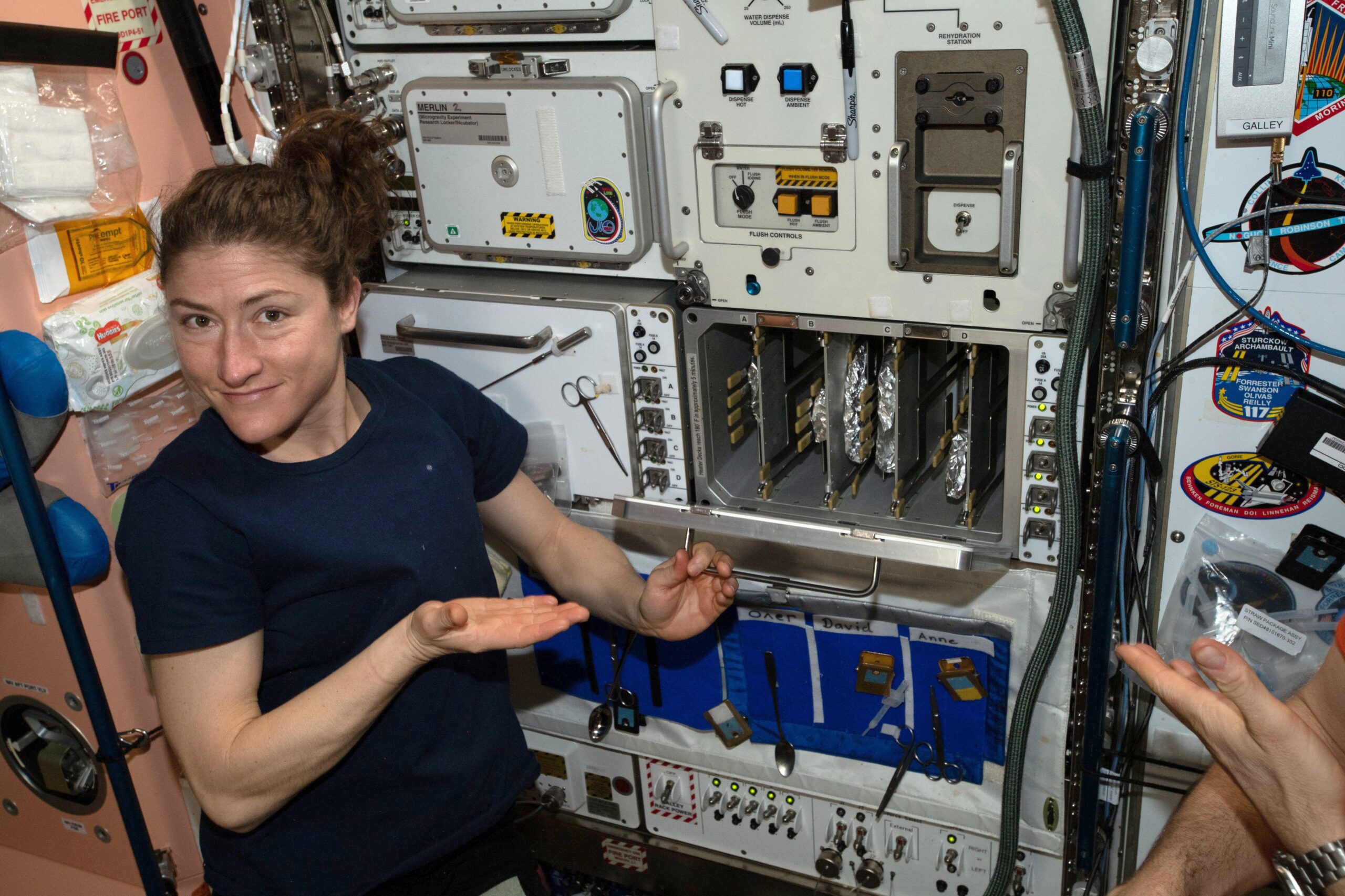 Smiling astronaut gestures toward a galley rack with foil-wrapped packets inside a space station module, with tools, cables and mission patches around.